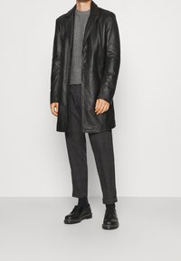 Černý kožený kabát s klopovým límcem, pod ním texturovaný šedý svetr a vzorované tmavé kalhoty, doplněné černými šněrovacími botami.