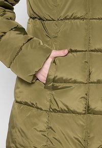 Chaqueta de plumas verde oliva con secciones acolchadas y un bolsillo lateral. La tela tiene una textura suave y brillante con una silueta ajustada.