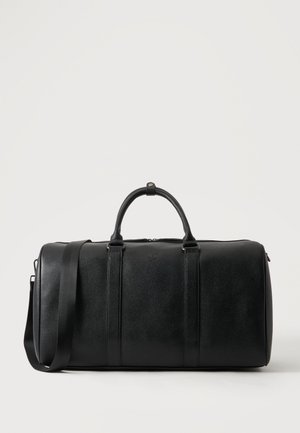 Sac de voyage en cuir noir avec deux poignées et une bandoulière réglable sur un fond clair uni.