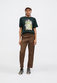 T-shirt vert foncé avec impression graphique, pantalon marron et baskets grises ; coupe droite avec une coupe décontractée et une casquette texturée.