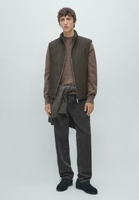 Ciemnozielona kamizelka bez rękawów na brązowym swetrze, zestawiona z ciemnoszarymi jeansami i czarnymi butami. Prosty design, miękka tekstura, casualowy krój.