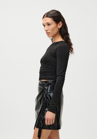 Fekete hosszú ujjú felső, oldalán ráncolt részletekkel, magasan ültetett, fényes fekete bőr rakott szoknyával, amelynek megkötős záródása van.