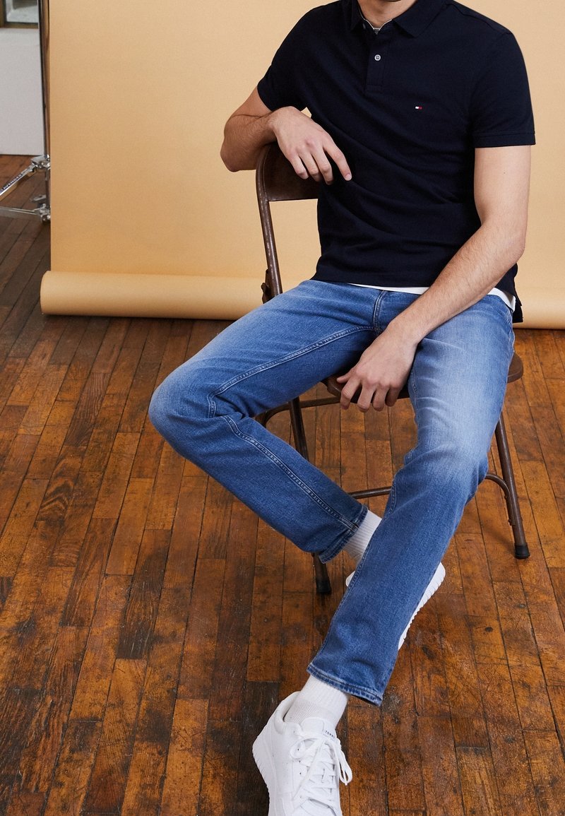 Polo shirt bleu marine avec un petit logo, jeans en denim bleu et baskets blanches. Assis sur une chaise en bois contre un fond beige.
