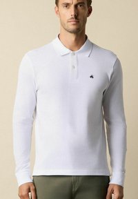 Biała, długosleeve polo wykonana z miękkiego materiału, z kołnierzykiem oraz małym ciemnym logo na piersi. Prosty design bez wzorów.