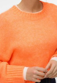 Personne portant un pull en tricot orange vif avec col et poignets côtelés, mains jointes devant, par-dessus une chemise blanche à manches longues.
