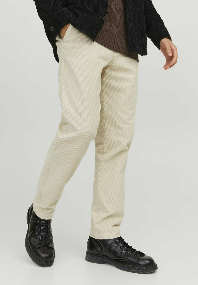 Beige corduroy broek met verticale ribbels, gecombineerd met zwarte veterschoenen. De outfit heeft een donkere textuurjack en een gebreide trui.