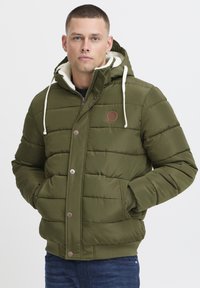 Olivgrüne Steppjacke mit einer gefälschten Lammfell-futterten Kapuze, gerippten Bündchen und Fronttaschen. Verfügt über braune Druckknöpfe und ein Logo-Patch.