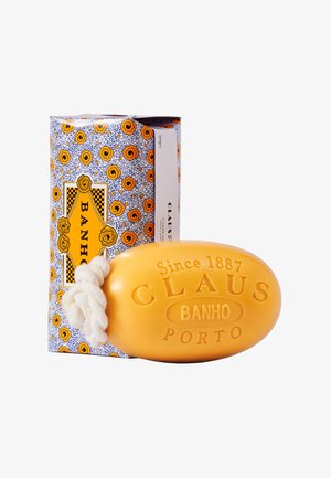 Ovaler gelber Seifenblock mit weißem Seil und eingeprägtem "CLAUS BANHO PORTO Seit 1887" neben einer dekorativen Schachtel mit orangen Rosenmustern.