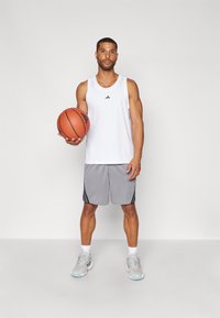 Camisola de malha branca sem mangas com logótipo preto, calções cinzentos com riscas pretas e sapatilhas cinzentas. A segurar uma bola de basquete. Fundo simples.