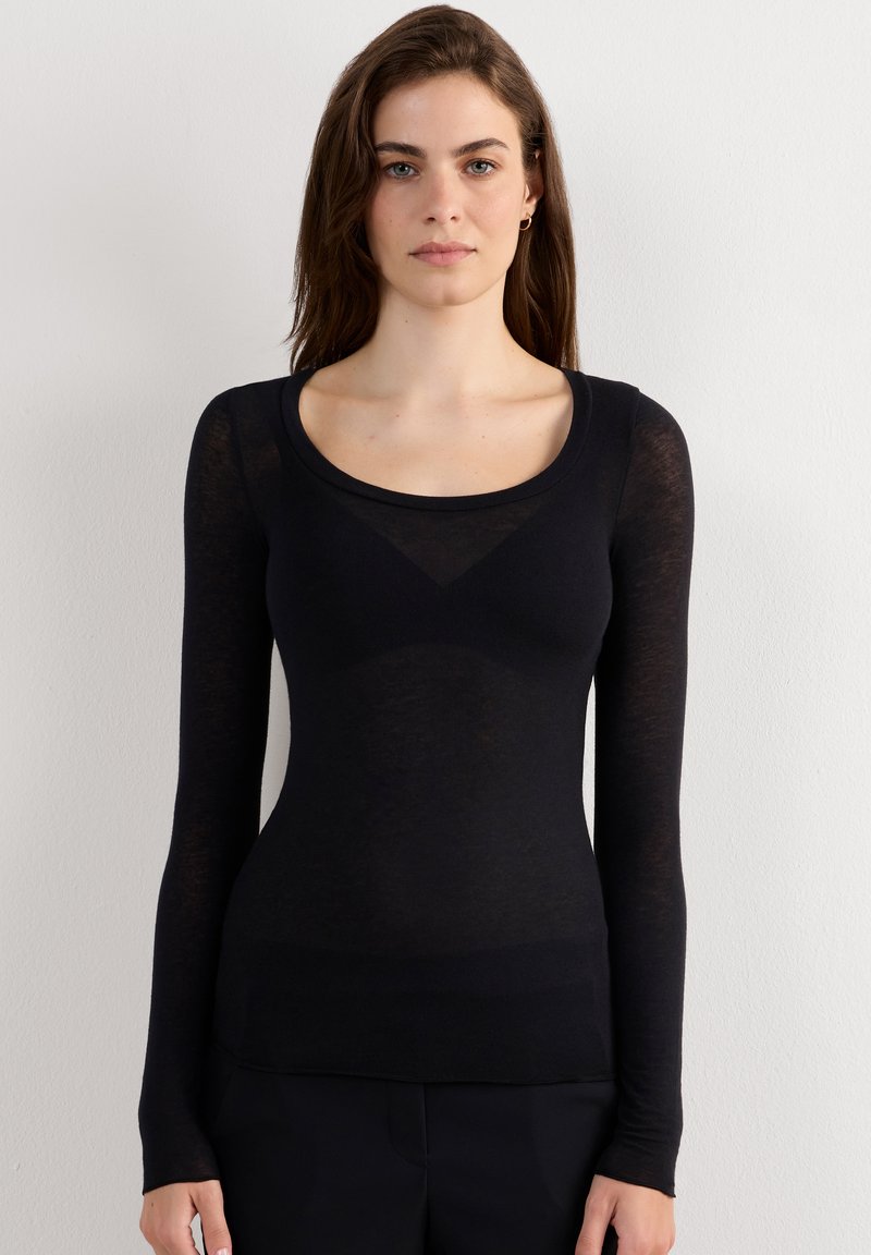 Jonge vrouw met lang bruin haar, gekleed in een aansluitende zwarte top met lange mouwen en zwarte broek, staand tegen een effen witte muur.