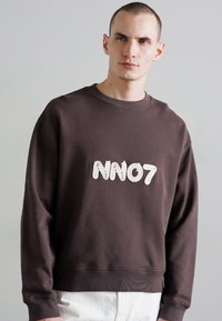 Sweat-shirt en coton brun avec texture côtelée, col rond et logo "NN07" brodé en blanc à l'avant. Le modèle porte un pantalon blanc.