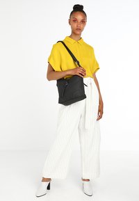 Sac à bandoulière en cuir noir avec une finition texturée, une sangle unique, une fermeture éclair et des détails de couture subtils, présenté sur un fond blanc.