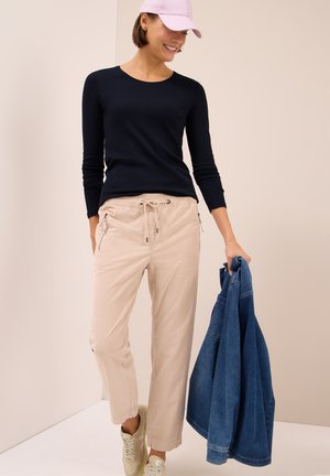 Eine Frau trägt ein schwarzes Langarmshirt, beige Hosen und helle Sneakers, hält in einer Hand eine blaue Jeansjacke und lächelt unter einer rosa Mütze.