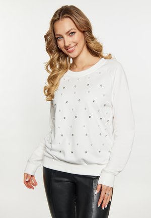 Kvinde med langt, bølget blond hår iført hvid sweatshirt med små skinnende pynt og sorte læderbukser, smilende til kameraet.