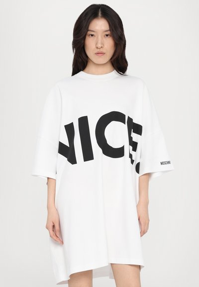 Nadrozmerné biele tričko-dress vyrobené z mäkkého materiálu. Obsahuje veľký čierny text "NICE" na prednej strane a logo "MOSCHINO" na rukáve.
