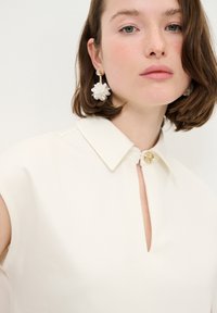 Cremfarbener Oberteil mit einem Kragen, Schlüssel-Halsauschnitt und Knopfverschluss. Besticht durch einen strukturierten Stoff und ein Model, das florale Tropfohrringe trägt.