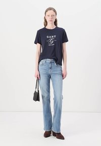 Marineblå T-shirt med hvid tekst, lyseblå jeans og mørkebrune hælsko. Desuden holdes en lille sort håndtaske i den ene hånd.