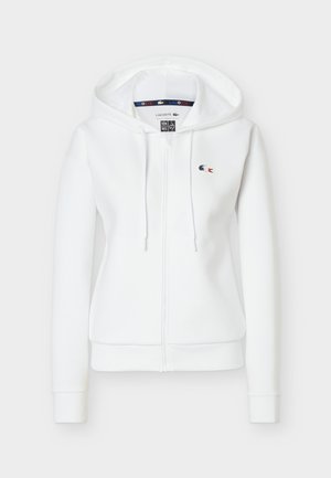 Valge tõmblukuga kapuutsiga dressipluus, millel on nöörid ja vasakul rinnal väike tikitud Lacoste'i logo.