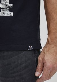 Marineblå bomullst-skjorte med hvit grafisk trykk. Har et sidemerke merket "FQ1924 ORIGINAL" og mørk grå jeans synlige.