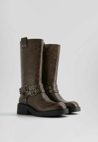 Hnědé kožené vysoké boty po kolena s širokým podpatkem, zdobené kovovými hřebíčky a přezkou na pásku kolem kotníku. Hladká textura.