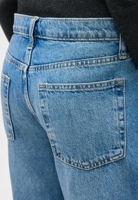 Κοντινό πλάνο στις πίσω τσέπες και τη μέση ενός ζευγαριού μπλε τζιν, φορεμένα με μαύρο τοπ, που δείχνει το ράψιμο και την υφή του υφάσματος.