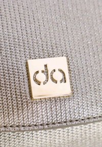 Bolso de tela tejida plateada con un acabado texturizado, que presenta una placa de logo dorada. Forma compacta con líneas limpias y detalle de costura visible.