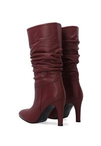 Pár uvolněných kožených bot burgundské barvy do poloviny lýtek, s špičatými špičkami a štíhlými vysokými podpatky na bílém pozadí.