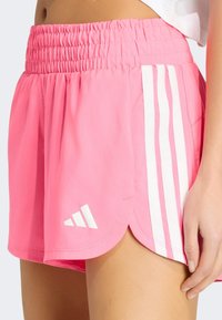 Shorts de sport roses avec une ceinture élastique, présentant des rayures verticales blanches sur les côtés et un logo blanc à l'ourlet.
