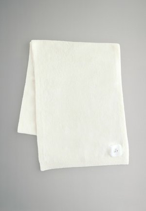 Hvid fleece tæppe med en blød tekstur, der har en ribbet kant og et lille mærke, der står "THE SET." Firkantet form på grå baggrund.