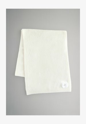 Bílá fleecová deka s jemnou texturou, zdobená žebrovaným okrajem a malou etiketou s nápisem "THE SET." Obdélníkový tvar na šedém pozadí.