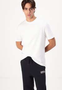 Weißes Baumwoll-T-Shirt mit kurzen Ärmeln und Rundhalsausschnitt, kombiniert mit dunkelblauen Jogginghosen, die ein Logo-Patch auf dem Oberschenkel haben.