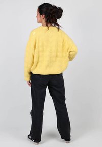 Un pull en maille jaune avec une coupe ample et un motif complexe, associé à un pantalon large noir et des baskets, vu de dos.