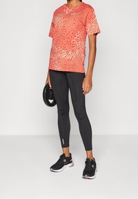 Punainen ja oranssi kuvioitu urheilupaita, yhdistetty mustiin leggingsseihin ja mustiin tennareihin. Asukokonaisuus sisältää kontrastoivia tekstuureja ja kerroksellisuutta.