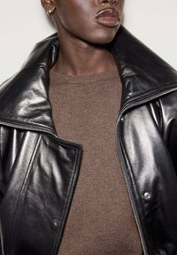 Chaqueta de cuero negra con cuello ancho, frontal con cremallera y cierre de botones, superpuesta sobre un suéter de punto marrón. Textura suave y ajuste relajado.