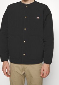 Person wearing a black quilted jacket with gold buttons and two front pockets, paired with beige pants against a plain background.