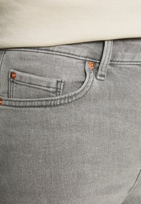 Szare dżinsy deniowe o gładkiej teksturze, z małą kieszonką z przodu i mosiężnymi nity na pasku.