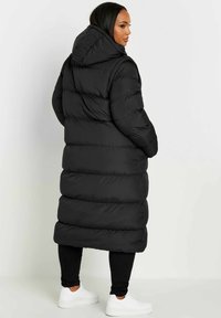 Svart puffjacka med en lång, vadderad design, huva och horisontell kviltad textur, bärs över svarta leggings och vita sneakers.