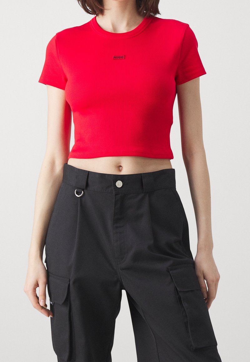 Personne portant un haut court rouge à manches courtes avec le logo "HUGO" et un pantalon cargo noir taille haute avec poches latérales et passants pour ceinture.