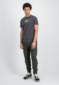 Grå bomulls t-skjorte med et neonlogo, kombinert med grønne cargo-bukser med flere lommer og en smal passform. Svarte og kremfargede sneakers.