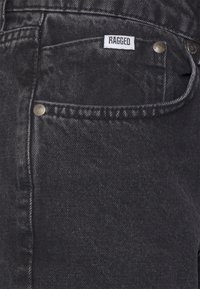 Jeans de mezclilla negra con un diseño clásico, etiqueta que dice "RAGGED" y botones de metal en los bolsillos frontales. Tejido texturizado.