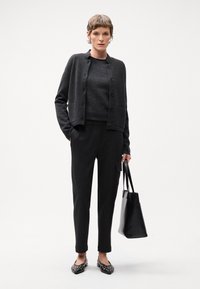Cardigan noir avec des boutons, pull gris, pantalon noir à rayures, et des chaussures plates noires avec des embellissements. Le modèle tient un sac fourre-tout noir.