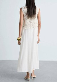 Maxikleid mit einem Spitzen-Crochet-Oberteil in Creme, das einen fließenden weißen Rock hat. Ein Armband mit grünen und gelben Akzenten wird am Handgelenk getragen.