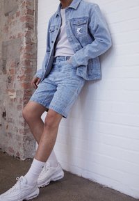 Lichtblauwe spijkerjas en shorts met ingewikkelde patronen. Witte T-shirt eronder, gecombineerd met witte sneakers en sokken.