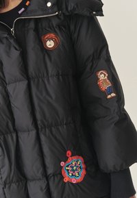 Juodas pūkinis švarkas pasižymi siuvinėta išvaizda, minkšta textura, siuvinėjimo detalėmis su lokiu ir vaiku bei spalvingu gėlių siuvinėjimu.