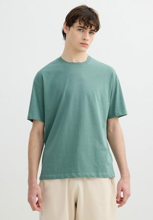 Joven con cabello castaño que lleva una camiseta verde lisa de manga corta y pantalones beige, de pie frente a un fondo blanco.