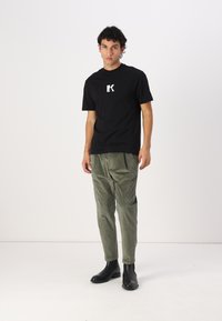 Zwart T-shirt met een wit logo, gecombineerd met olijfgroene corduroybroek en zwarte leren enkellaarsjes. De outfit heeft een relaxte pasvorm.