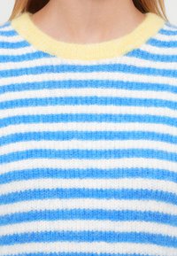 Monivärinen neulepusero, jossa vaakaraitoja vaaleansinisellä, tummansinisellä ja valkoisella. Keltainen resorinen pääntie. Pehmeä, pörröinen pinta.