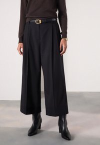 Sorte bukser med vide ben lavet af glat stof, med skarpe læg, en høj linning og et sort læderbælte med en guldlås.