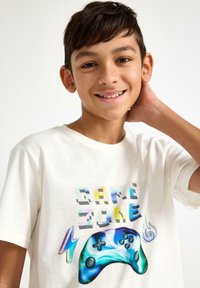 Witte katoenen t-shirt met de tekst "GAME ZONE" in kleurrijke holografische letters en een blauw gamecontroller-graphic met bliksem- en vlamaccenten.