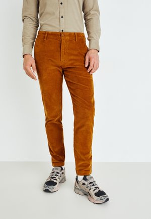 Mand iført rustfarvede kordbukser, beige skjorte med knapper, hvide sokker og sorte og grå sneakers, stående mod en ensfarvet baggrund.
